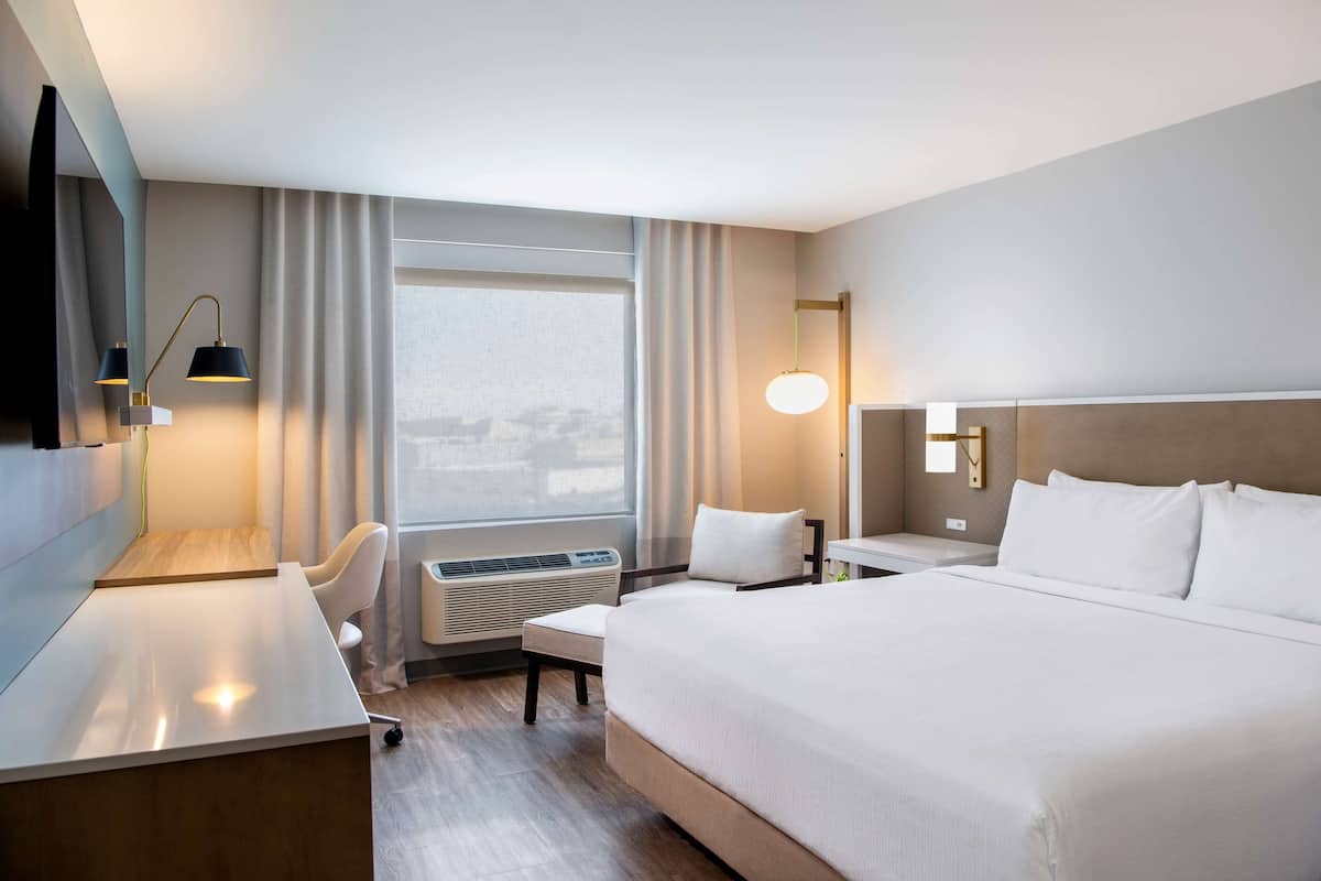 Una habitación de hotel con una cama grande, un escritorio, una silla, un televisor, una ventana con cortinas y una lámpara fijada a la pared.