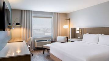 Una habitación de hotel con una cama grande, un escritorio, una silla, un televisor, una ventana con cortinas y una lámpara fijada a la pared.