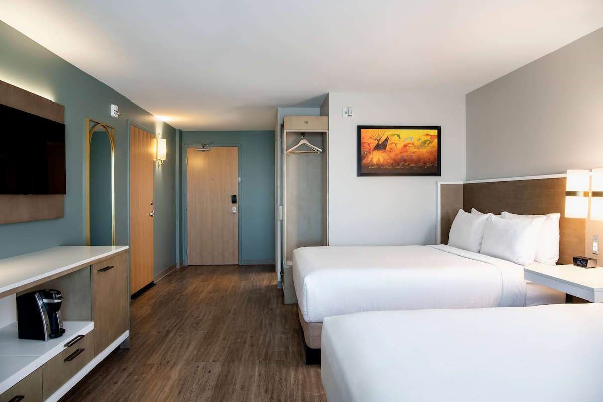 Habitación de hotel con dos camas, un armario, un televisor, un cuadro y una mesita de noche con lámpara.