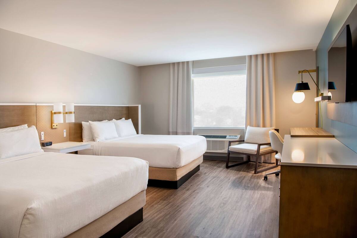 Habitación de hotel con dos camas, un escritorio, una silla, un televisor y un ventanal grande con cortinas.