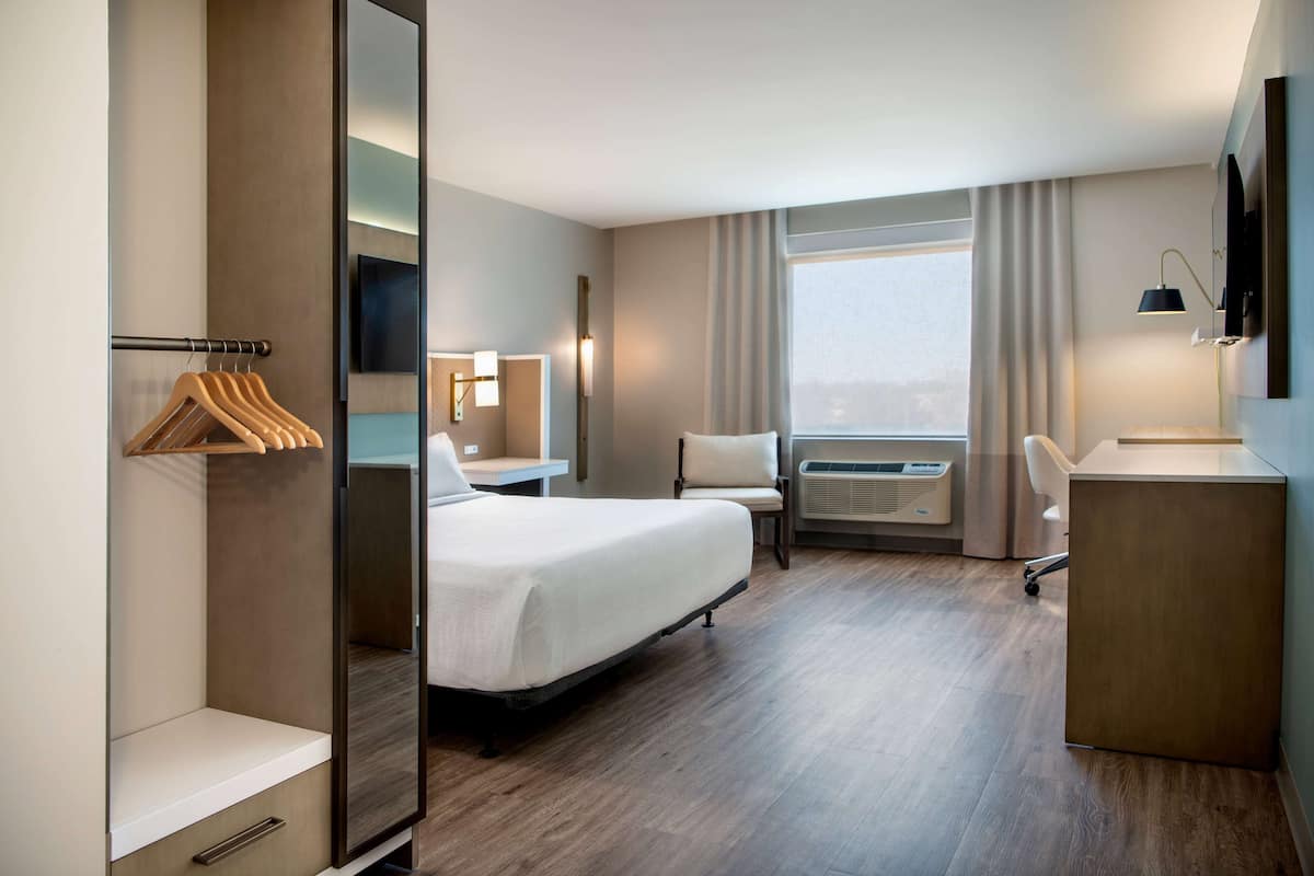 Habitación de hotel con cama, escritorio, silla, armario y ventana grande.