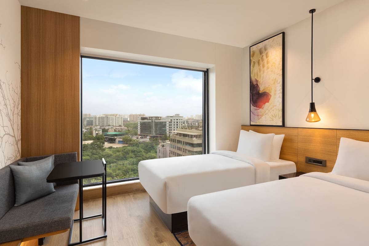 Ein Hotelzimmer mit zwei Betten, einem Schlafsofa, einem kleinen Tisch und einem großen Fenster mit Blick auf die Stadt.