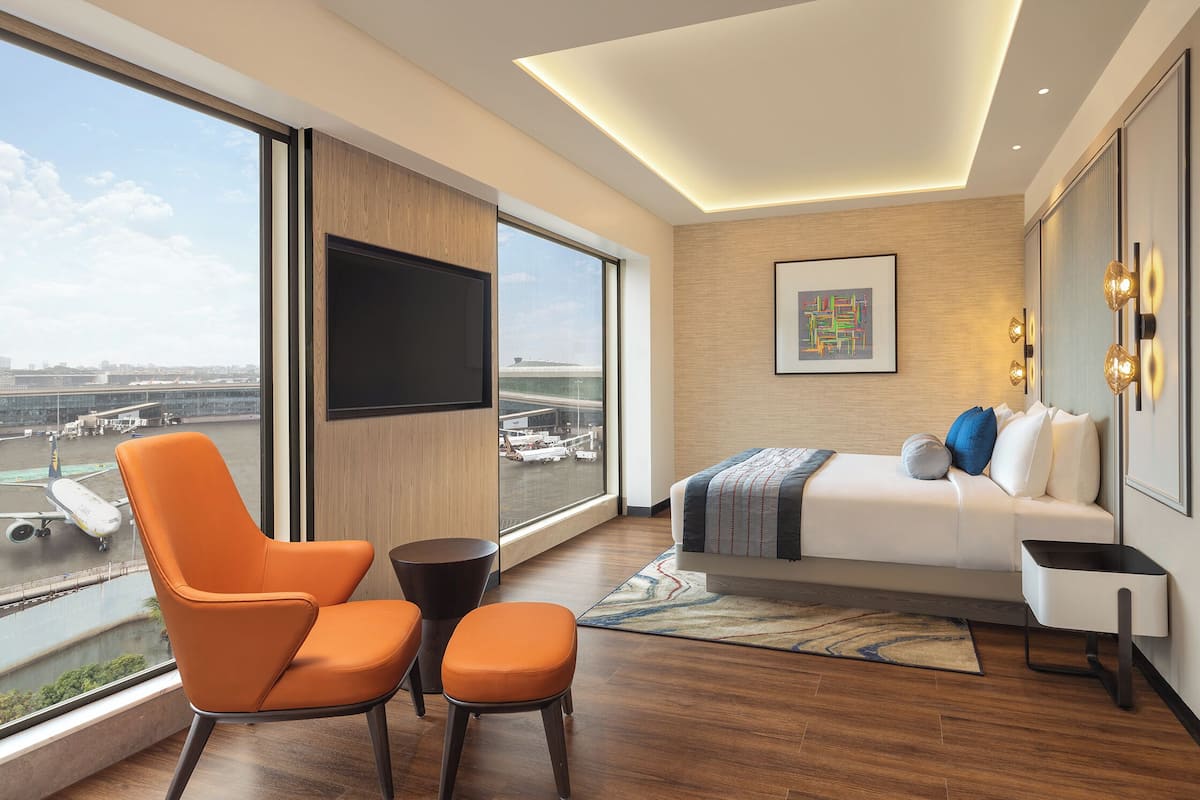 Ein modernes Hotelzimmer mit großem Fenster, einem Bett, einem Fernseher und Blick auf einen Flughafen.