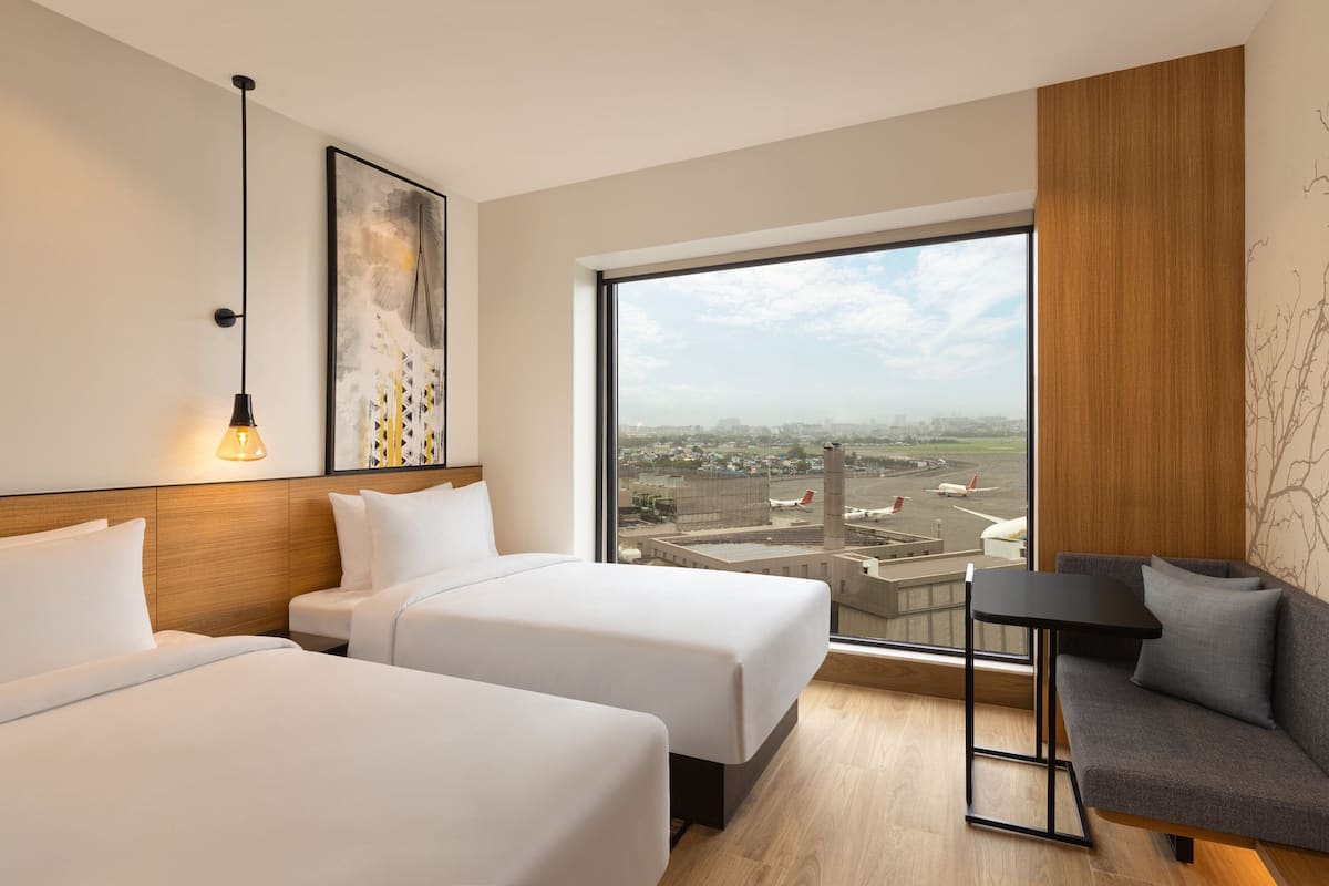 Ein Hotelzimmer mit zwei Betten, einem Sofa, einem kleinen Tisch, einem großen Fenster mit Stadtblick und einer Hängelampe.