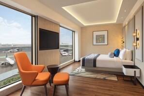 Ein modernes Hotelzimmer mit großem Fenster, einem Bett, einem Fernseher und Blick auf einen Flughafen.