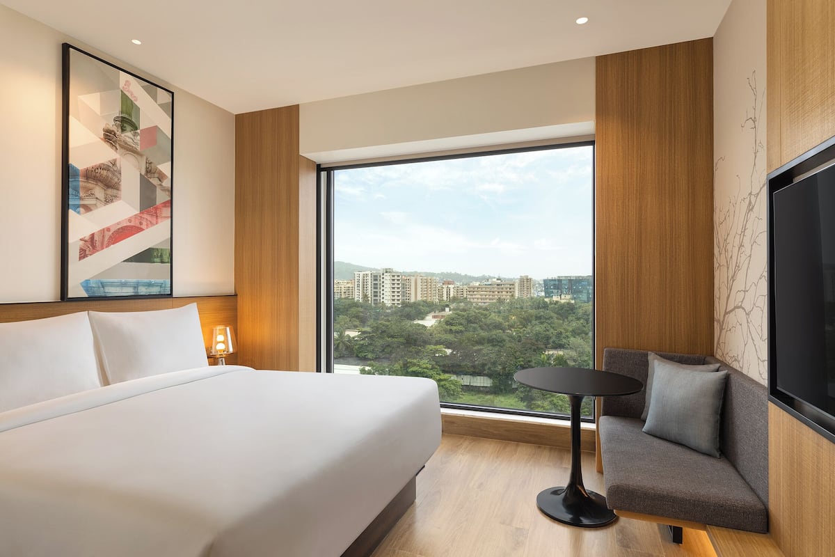 Ein Hotelzimmer mit einem großen Bett, einer Couch, einem kleinen Tisch, einem Fernseher und einem großen Fenster mit Blick auf die Stadt.