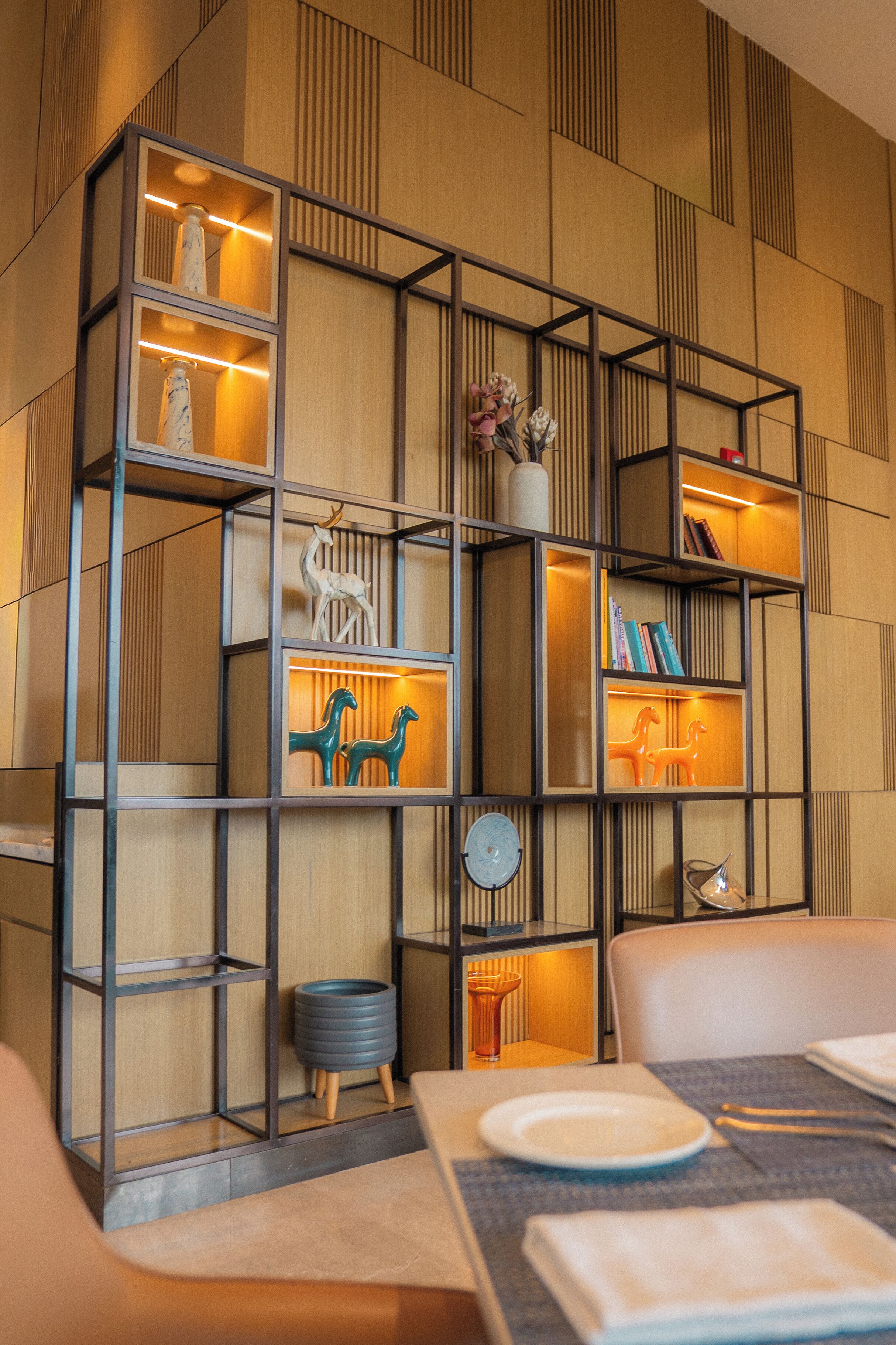 A modern interior with a wooden wall panel, a metal shelving unit with decorative items, and a dining area with a table set for two.