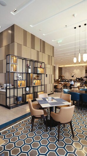 Eine moderne Hotelhalle mit einem geometrisch gemusterten Boden, einer Sitzecke mit Tischen und Stühlen sowie einer dekorativen Regaleinrichtung mit verschiedenen Gegenständen.