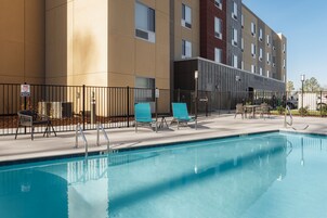 Piscina con sillones de descanso y una cerca, junto a un edificio moderno de varios pisos.