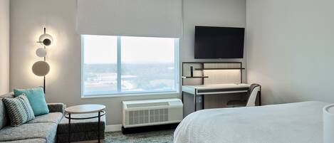 A hotel room with a bed, a sofa, a desk, a flat-screen TV, a window with a view, and a wall-mounted air conditioner.