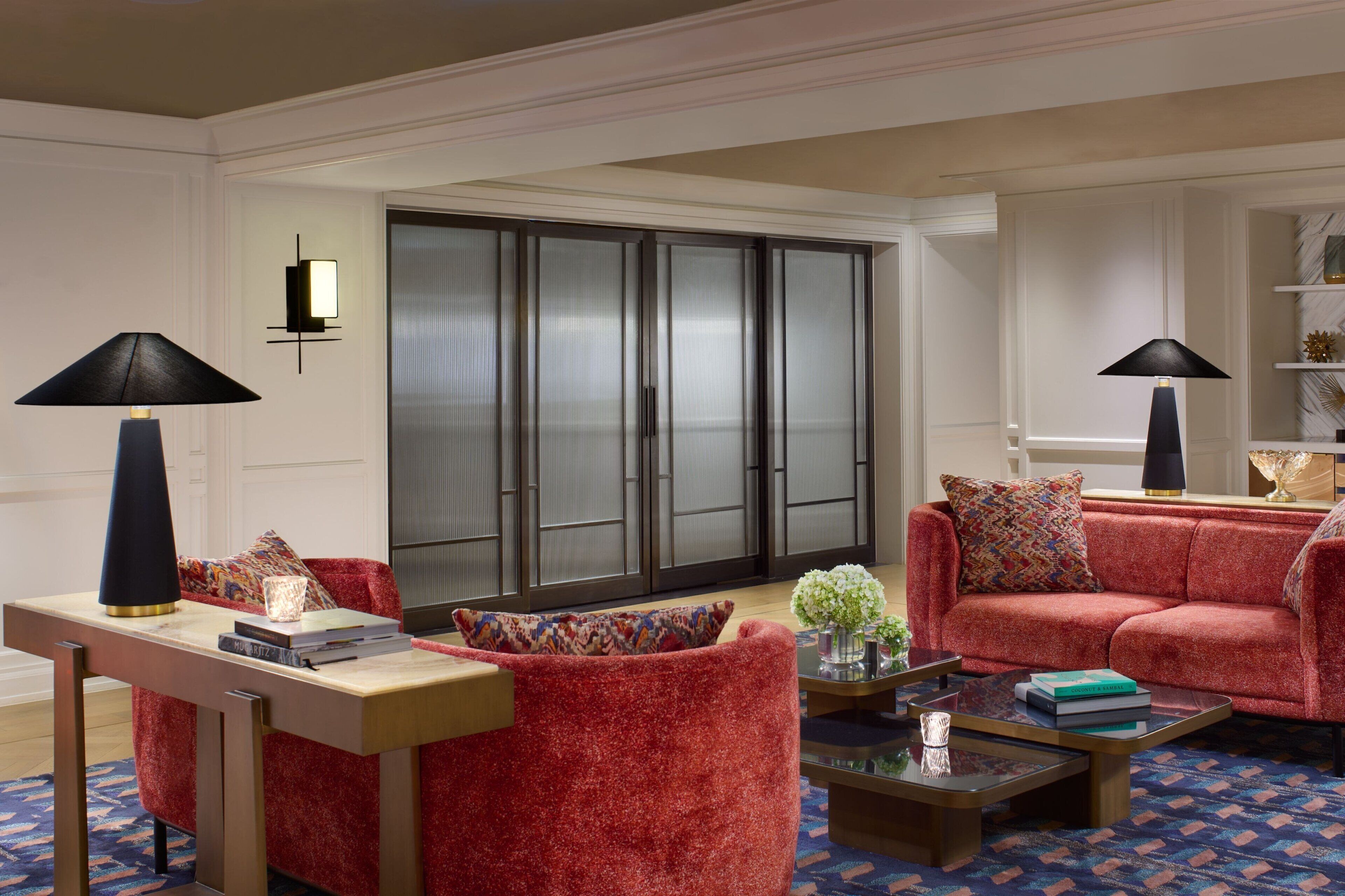 A modern living room with a red sofa, a coffee table, and two lamps. There is a bookshelf with decorative items and a sliding glass door in the background.
