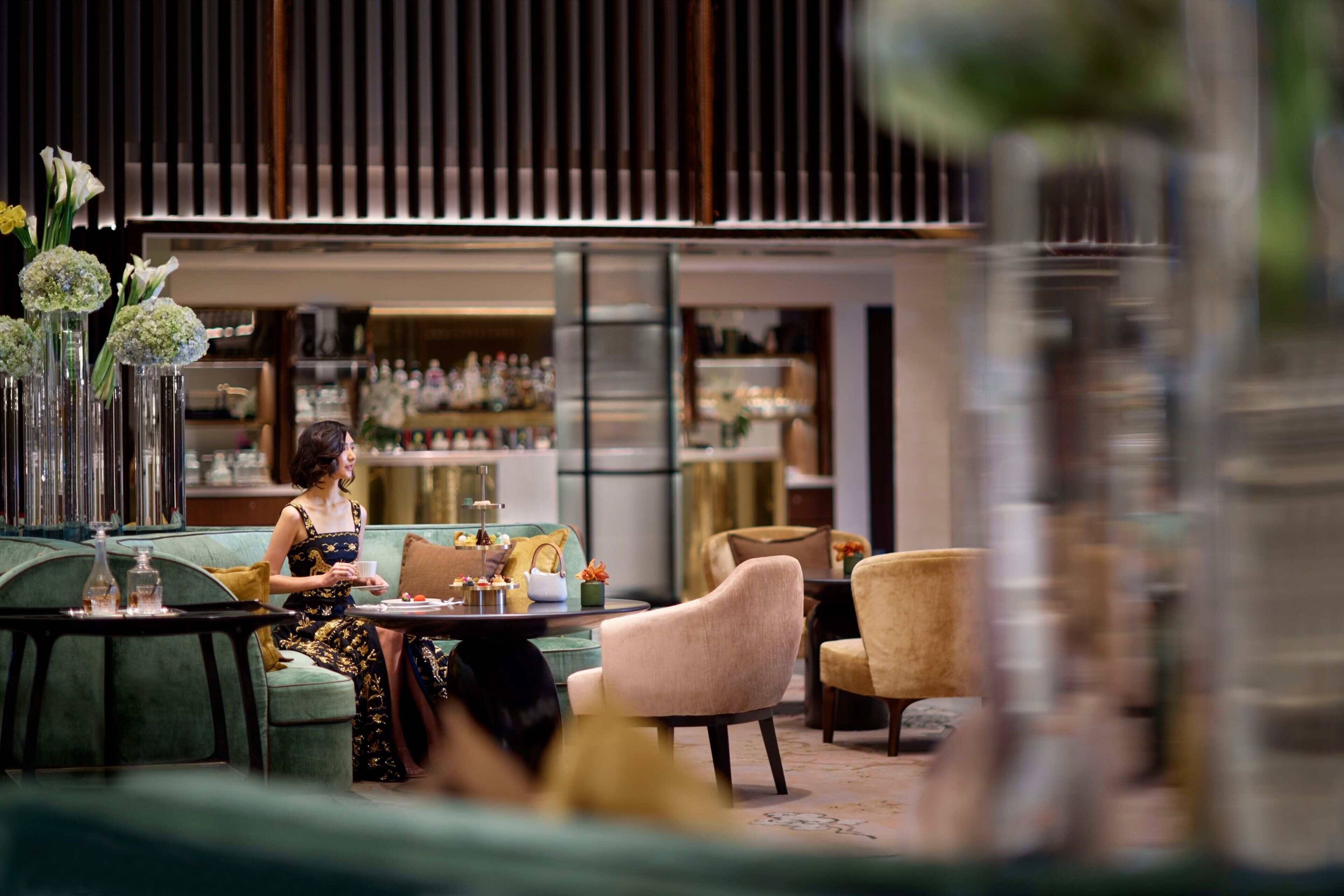 Een vrouw zit op een groene bank in een modern interieur, met een barretage op de achtergrond.