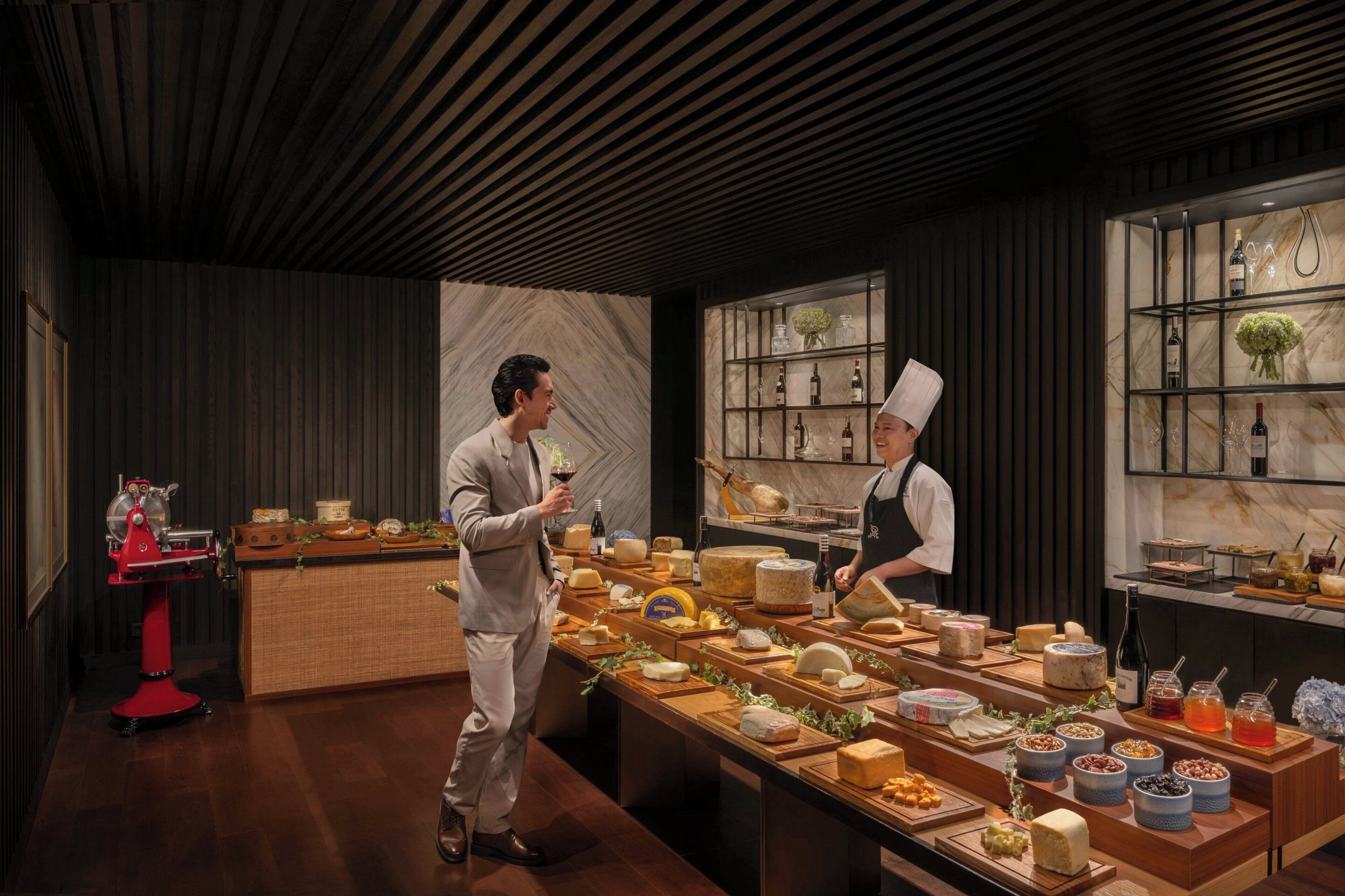 A chef and a guest in a modern kitchen with a marble backsplash, wooden counter, and various cheeses and wines displayed.