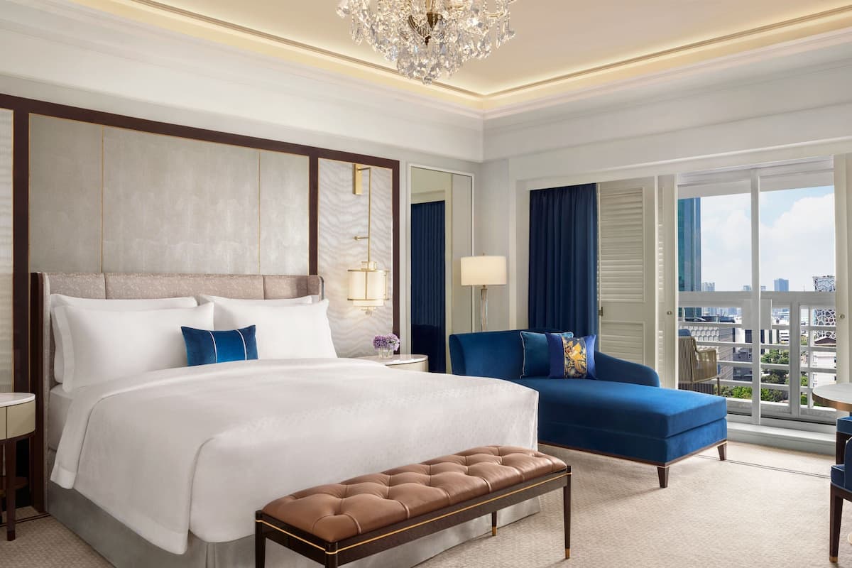 A hotel room with a large bed, a bench, a blue sofa, a chandelier, and a city view through the windows.