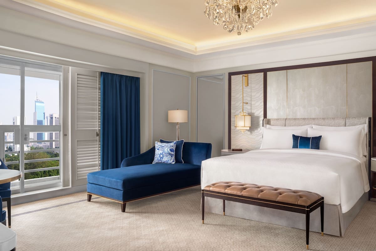 A hotel room with a large bed, a sofa, a bench, a desk, and a view of the cityscape.