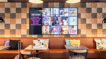 A modern lounge area with a large wall-mounted TV displaying various shows, a leather sofa, and a small table with a keyboard and a drink.