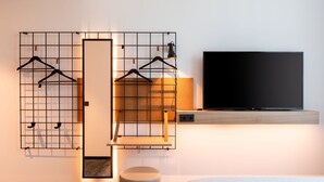 Una habitación de hotel moderna con un televisor de pantalla plana grande montado en un estante de madera, un espejo en la pared y dos estantes negros con ropa colgada.