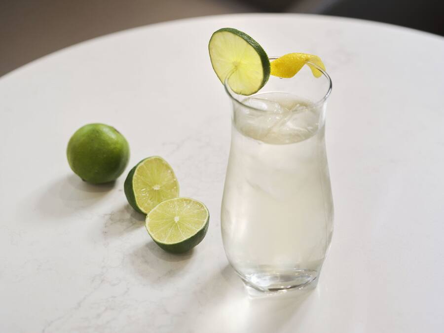 A glass of clear liquid with ice cubes, garnished with a lime slice and a yellow citrus peel, on a white marble surface.