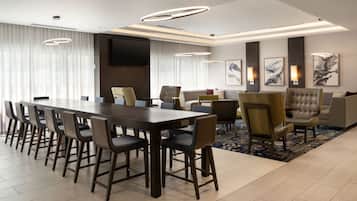 A modern hotel room with a large dining table, comfortable seating, a flat-screen TV, and artwork on the walls.