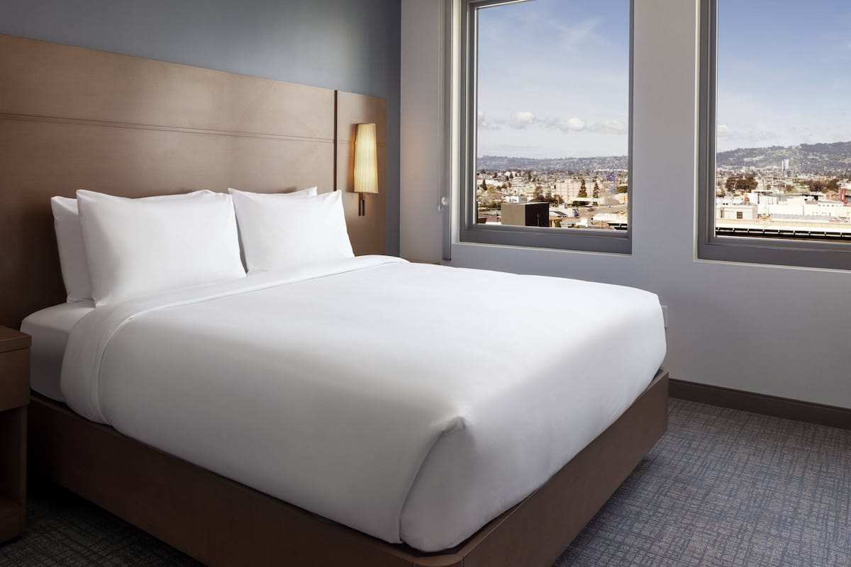 A hotel room with a large bed, a nightstand, a wall-mounted lamp, and a window with a city view.
