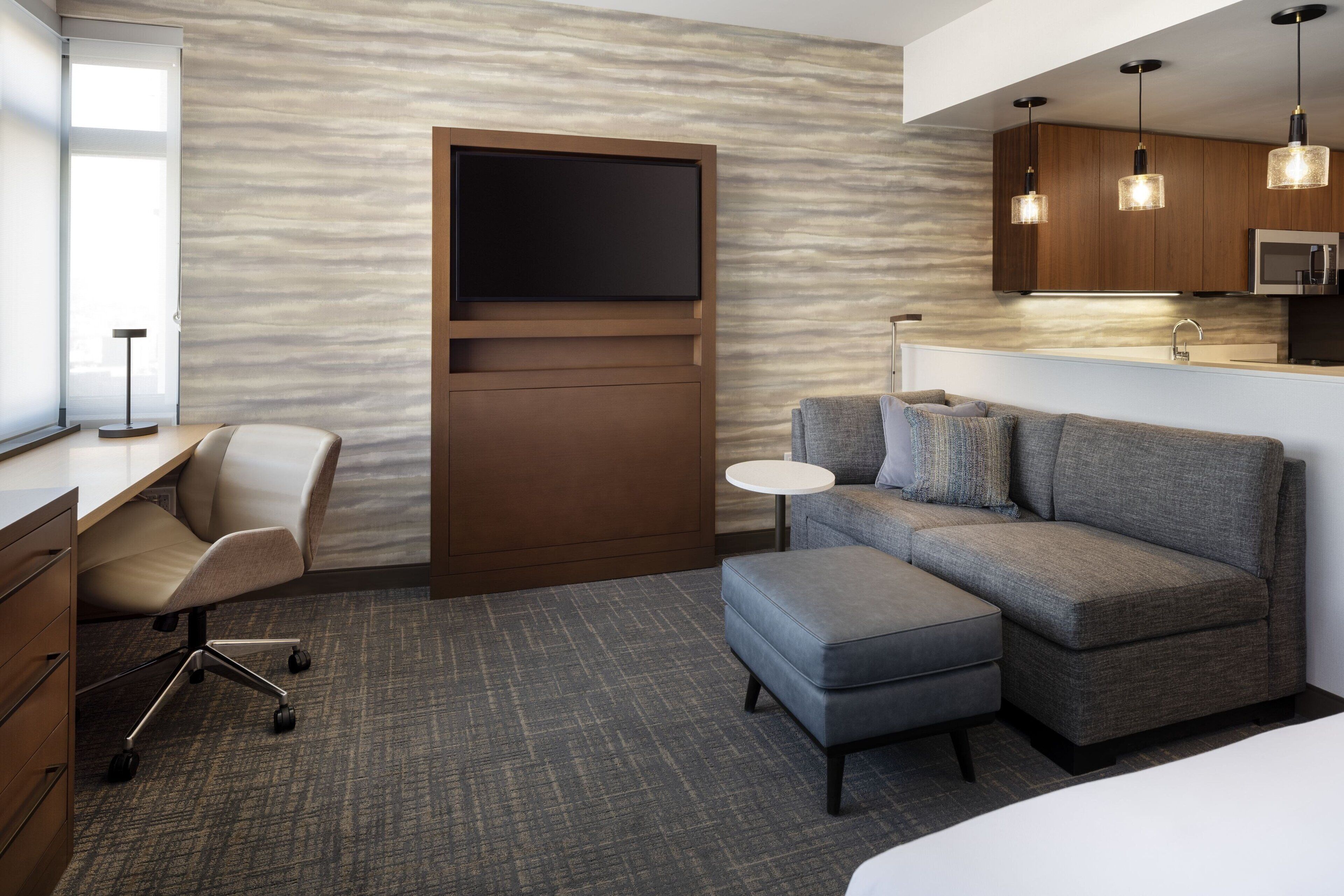 Una habitación de hotel moderna con un televisor de pantalla plana, un sofá, un reposapiés, un escritorio con una silla y una mesita redonda.