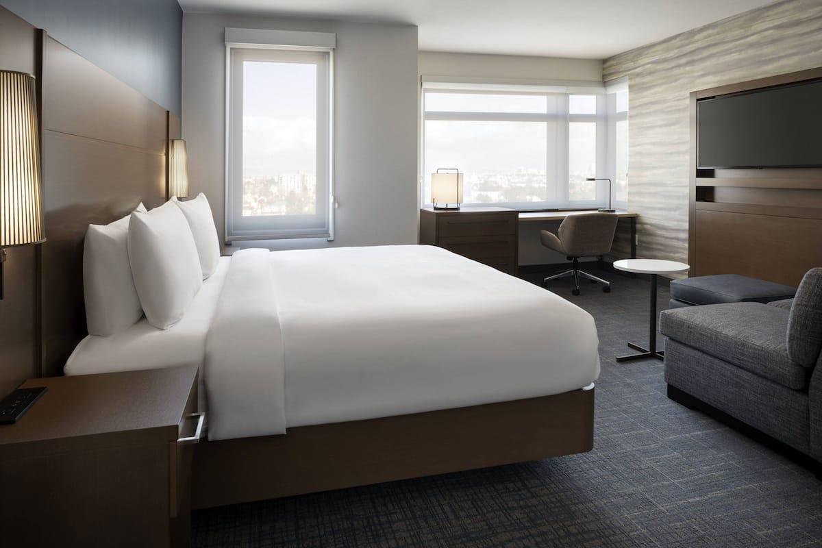 A hotel room with a large bed, a desk, a chair, a TV, and a view of the city.