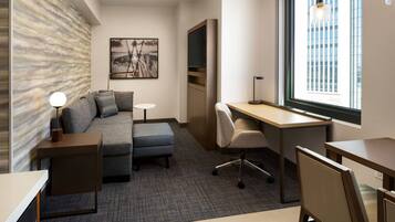 Una habitación de hotel moderna con un sofá gris, una mesita, un escritorio y una silla. Hay un cuadro en la pared y una ventana con persianas.