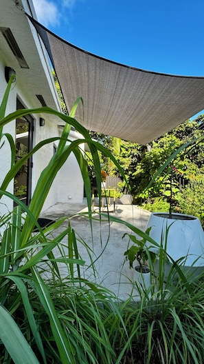 A modern outdoor patio area with a large shade sail, a concrete floor, and lush greenery.