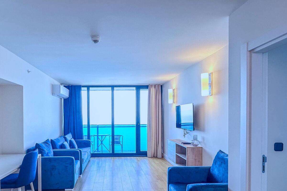Ein modernes Hotelzimmer mit Balkon, einer Sitzecke mit blauen Stühlen, einem Flachbildfernseher und einem Holznachttisch.