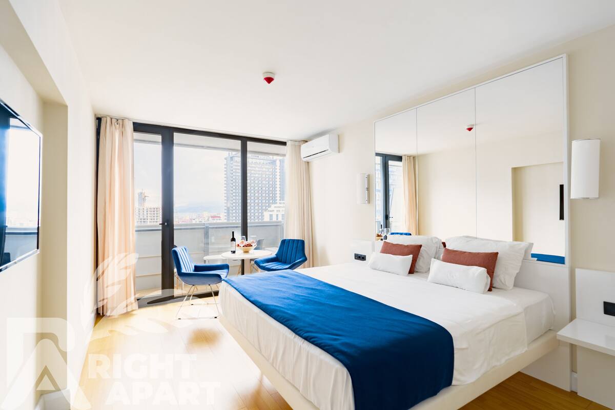 Ein modernes Hotelzimmer mit einem großen Bett, einem Schreibtisch, einem Sessel und Blick auf die Stadt durch große Fenster.