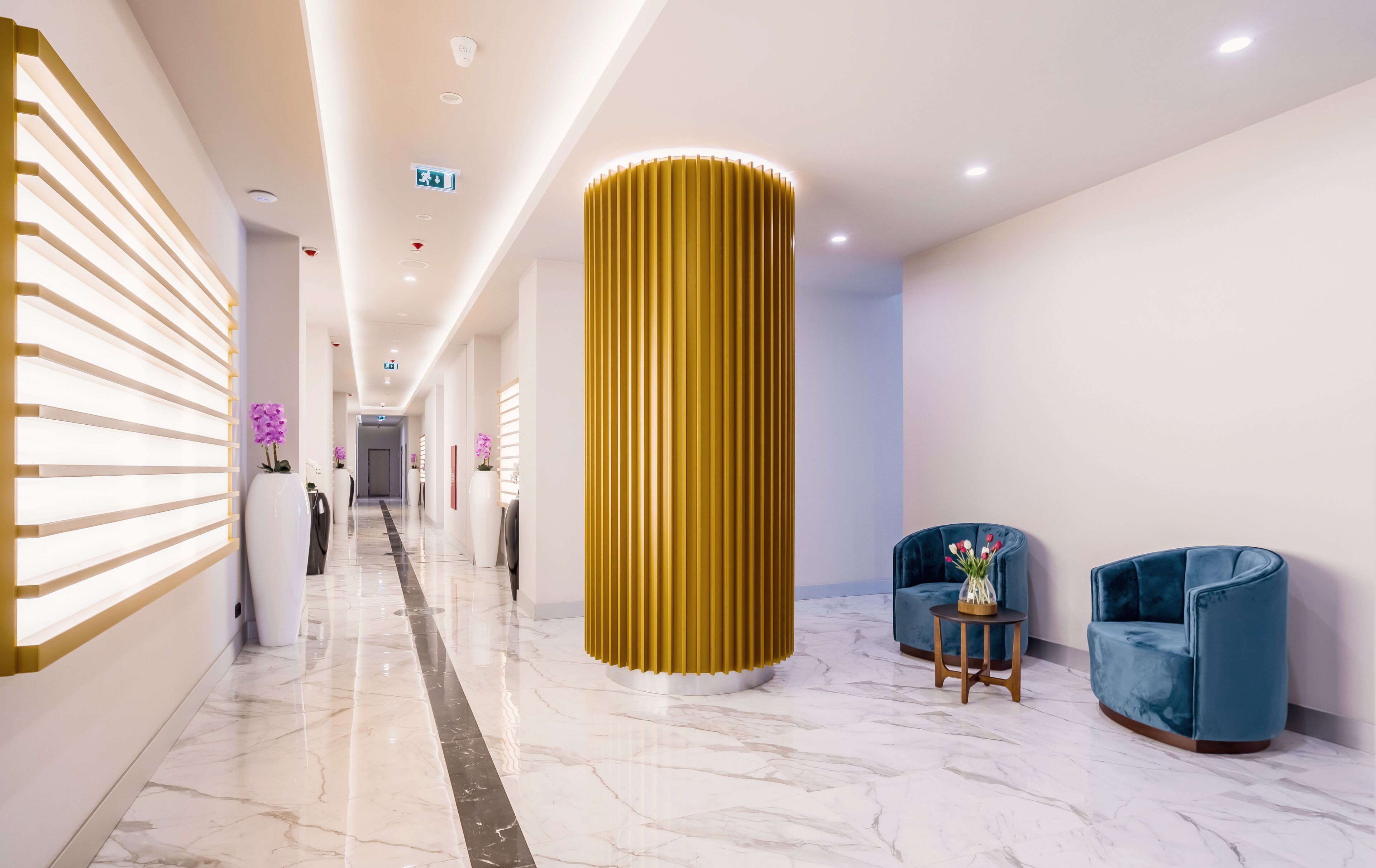 Un hall d’hôtel moderne avec un sol en marbre, une grande colonne dorée et des fauteuils bleus recouverts de tissu.