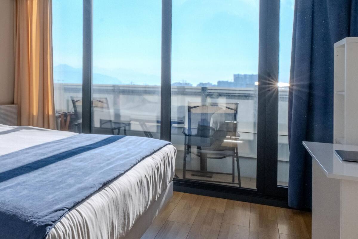 Ein Hotelzimmer mit einem großen Bett, einem Schreibtisch, einem Sessel und Blick auf die Stadt durch große Fenster.
