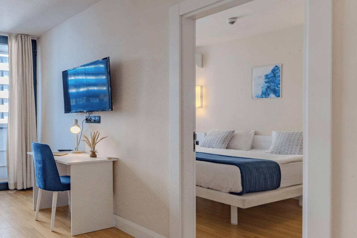 Ein modernes Hotelzimmer mit einem Flachbildfernseher, einem Schreibtisch mit Stuhl, einem Bett mit blauer Decke und einem Gemälde an der Wand.