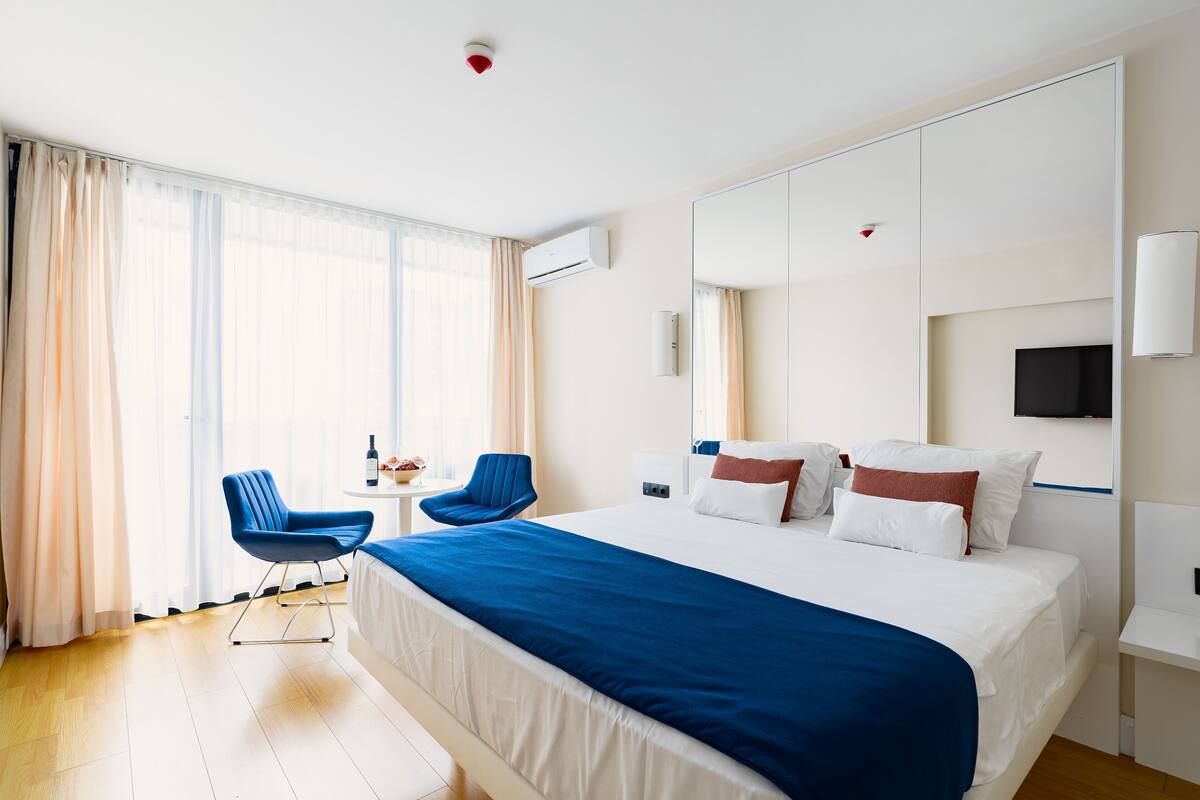Ein Hotelzimmer mit einem großen Bett, zwei blauen Stühlen, einem kleinen Tisch mit einer Flasche und einem Fernseher an der Wand.