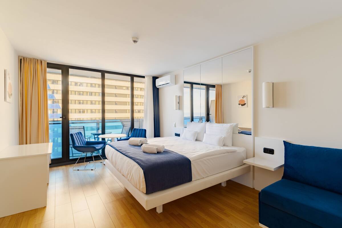 Ein modernes Hotelzimmer mit einem großen Bett, einem Schreibtisch, einem blauen Sofa und einem Balkon mit Blick auf Gebäude.