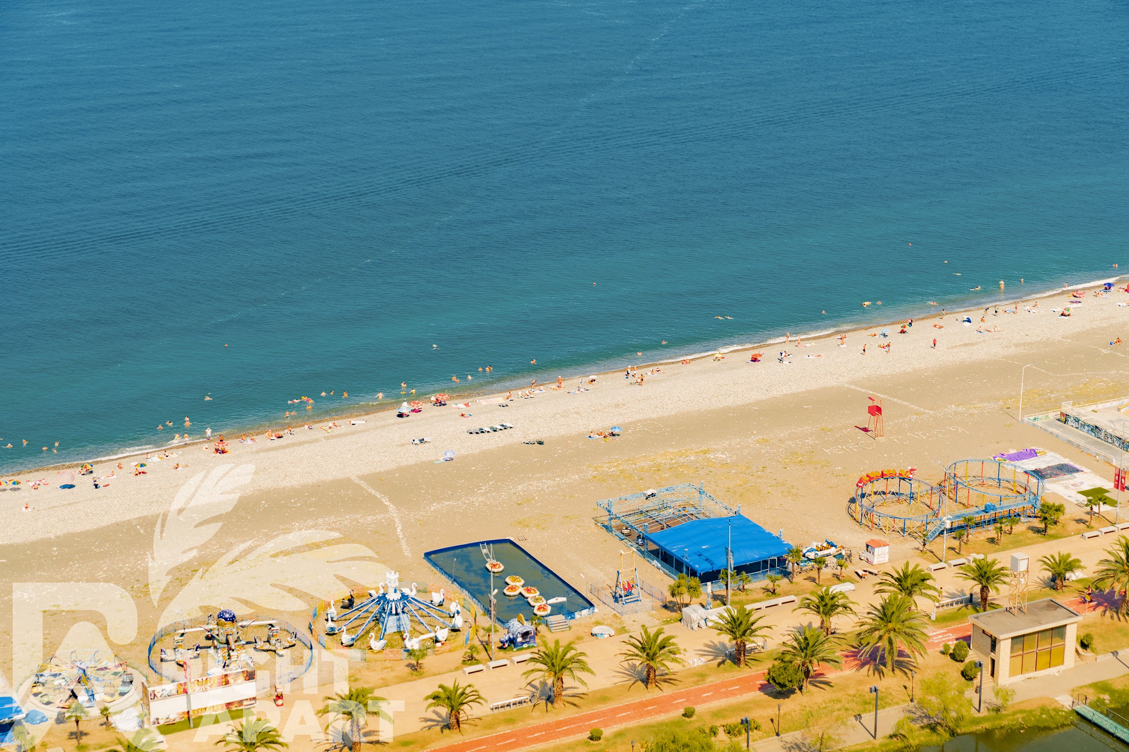 An aerial view of a beach with a swimming pool, palm trees, and a beachside restaurant.