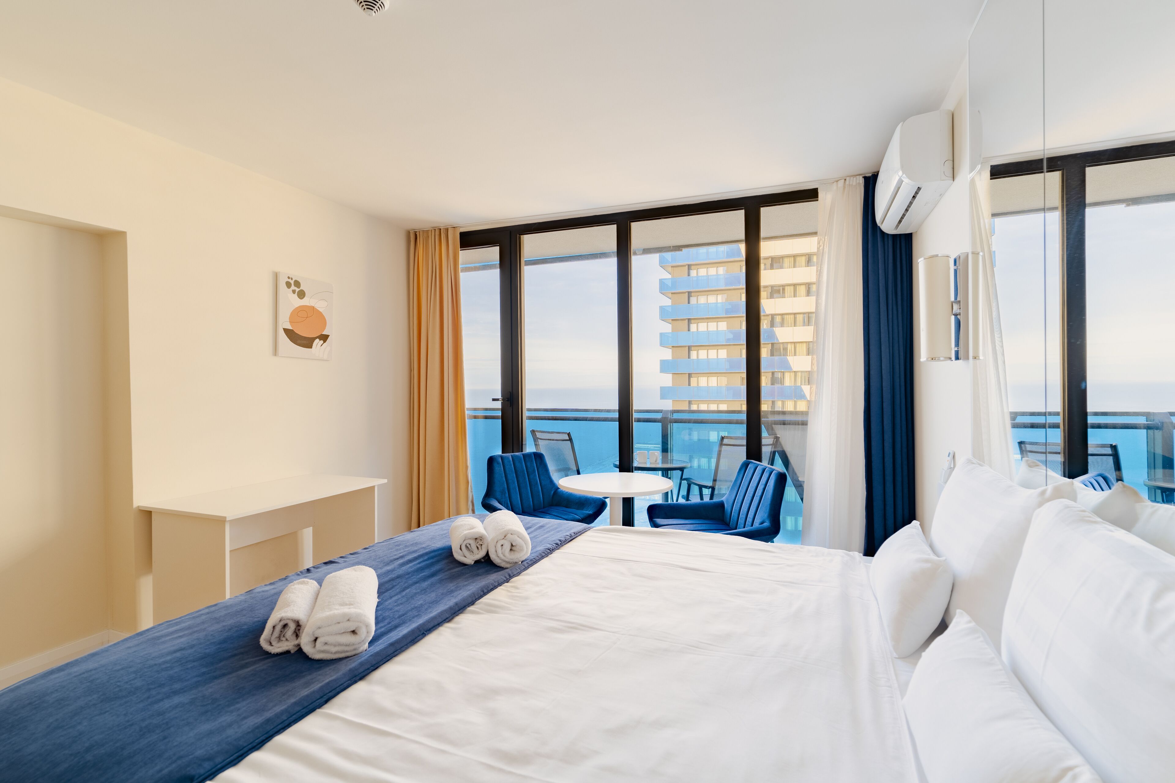 Een hotelkamer met een bed, een bureau, een stoel en uitzicht op zee en gebouwen.