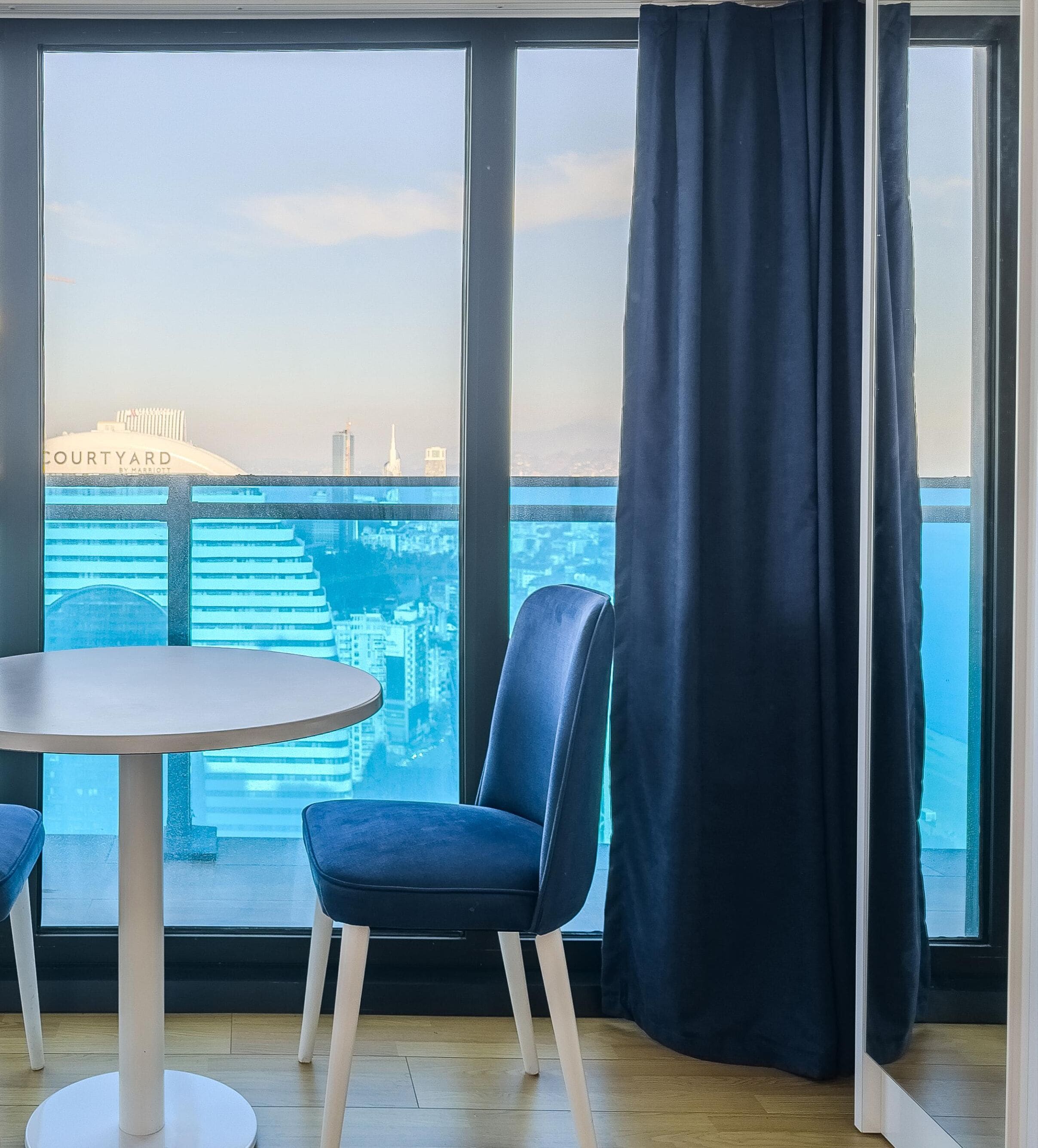 A room with a large window, a round table, a blue chair, and a dark curtain.