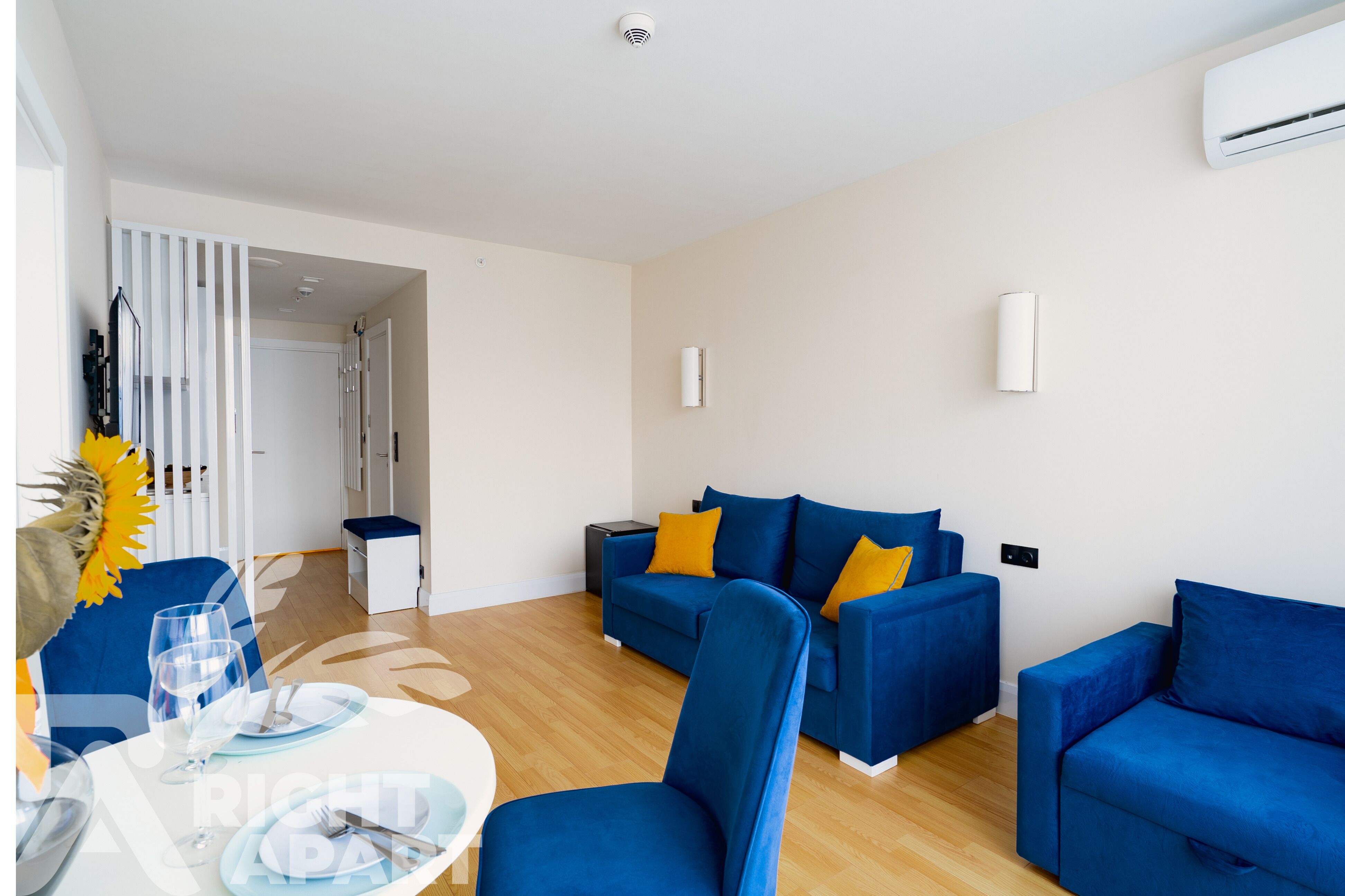 Una sala de estar moderna con sofás azules, una mesa de comedor con platos y vasos, y un equipo de aire acondicionado blanco en la pared.