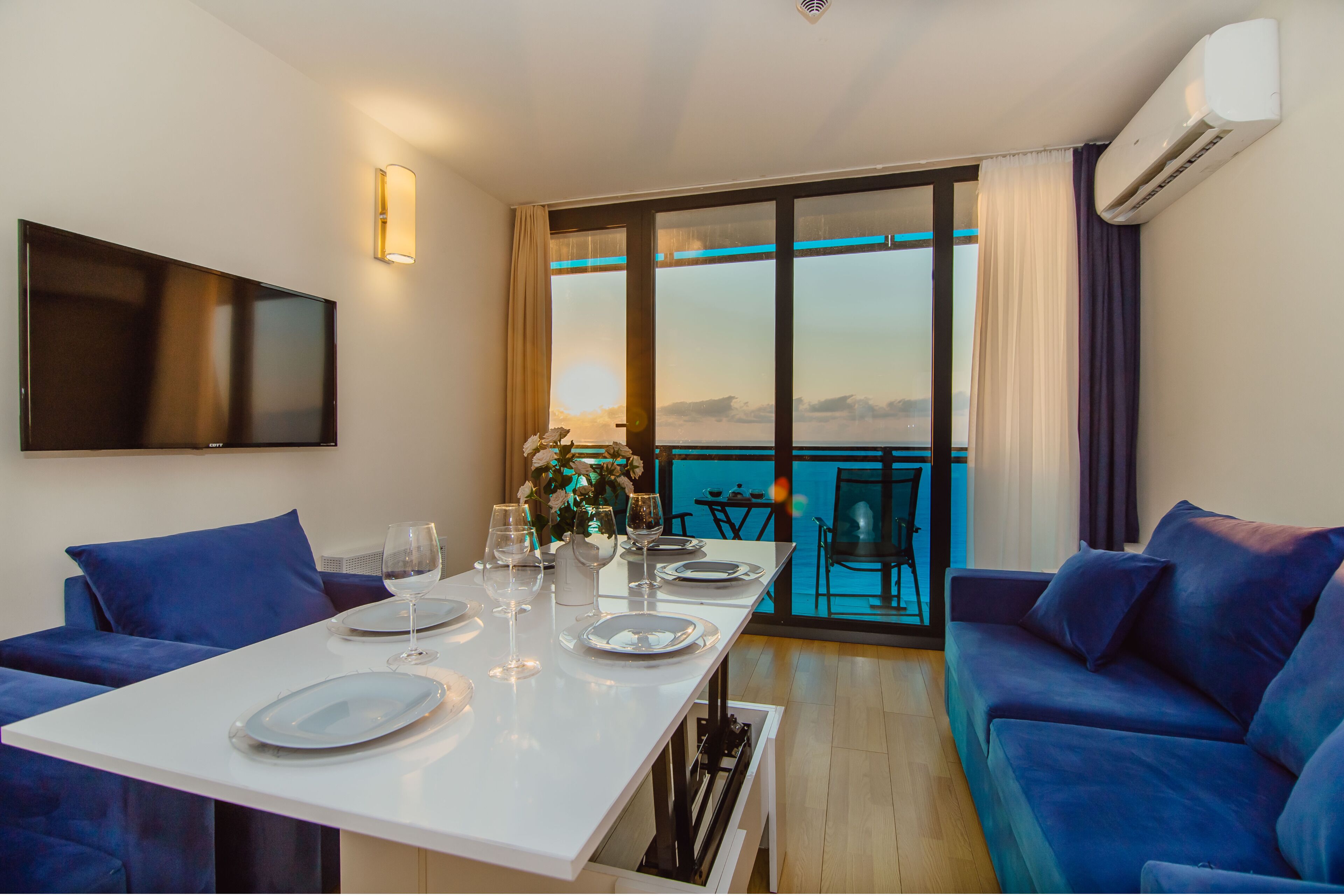 A dining area with a white table set for four, a sofa, and a TV mounted on the wall.
