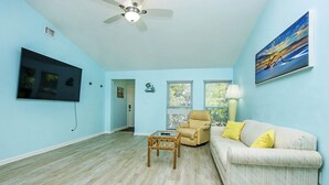 A living room with a ceiling fan, a flat-screen TV mounted on the wall, a sofa with yellow cushions, and a window with blinds. - Family Friendly, Great Pool and Easy Bowman's Beach Access (Sanibel)