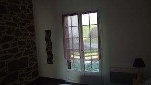 A bedroom with a stone wall, a bed with patterned bedding, a window with a view of greenery, and a bedside table with a lamp. - Countryside at the foot of Mont Saint Michel (Roz-sur-Couesnon)