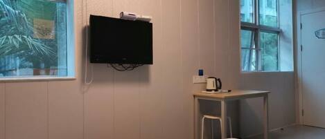 A hotel room with a bed, a small table with a kettle, a TV mounted on the wall, and a window with a view of trees.