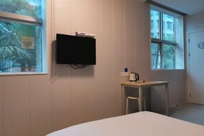 A hotel room with a bed, a small table with a kettle, a TV mounted on the wall, and a window with a view of trees. - Baishijie Hotel (Shantou)