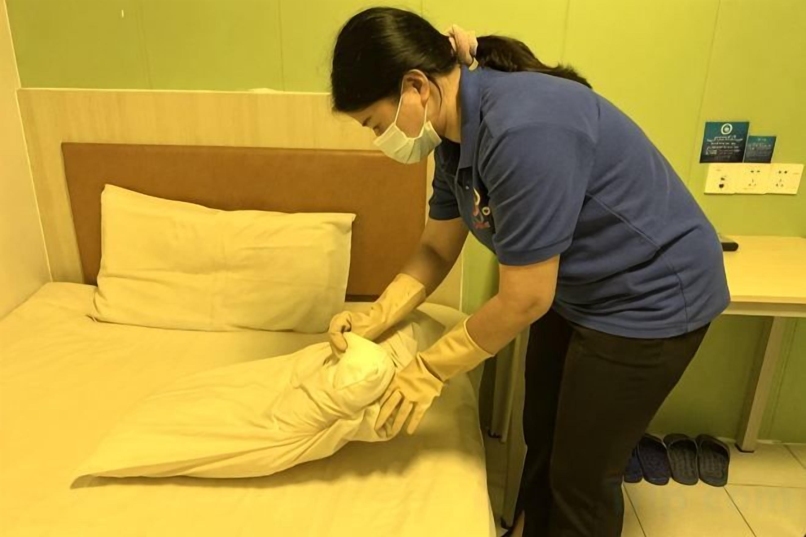 A person in a blue uniform adjusting a bedspread on a bed with a wooden headboard.