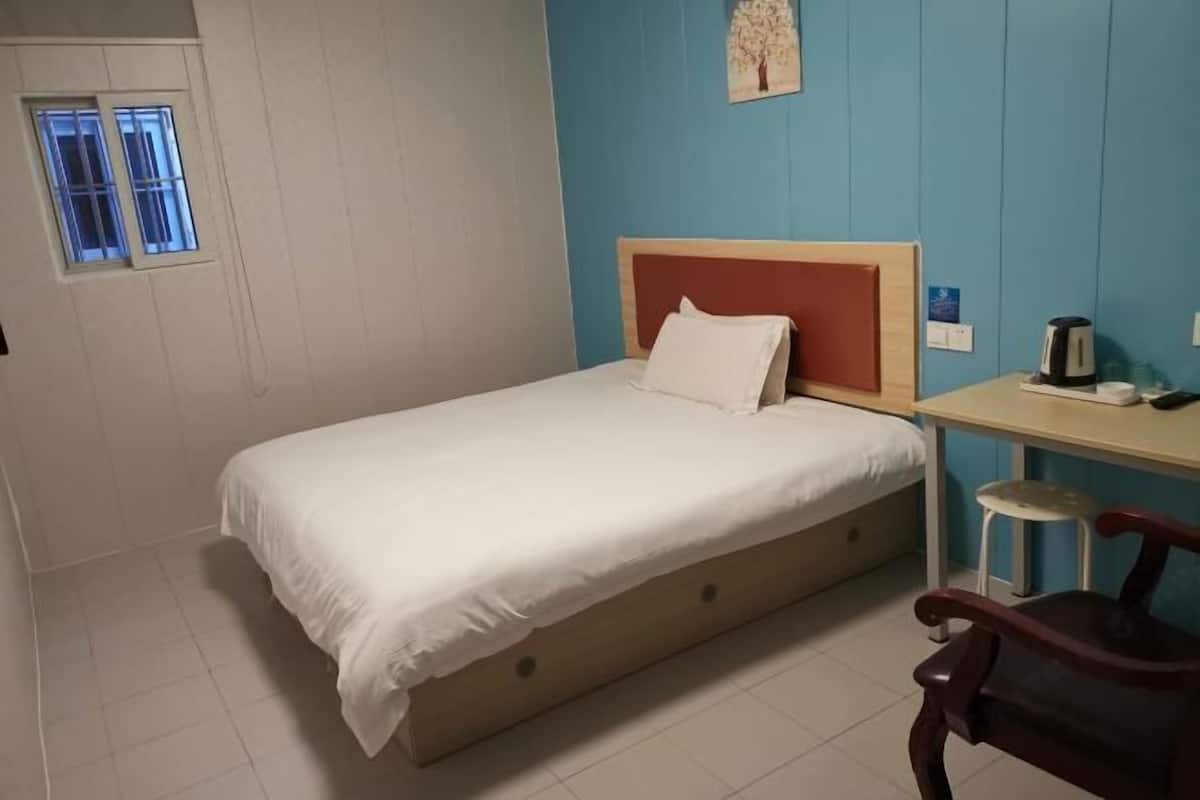 Uma cama de solteiro com cabeceira de madeira, uma pequena escrivaninha com cadeira e uma janela com grades.