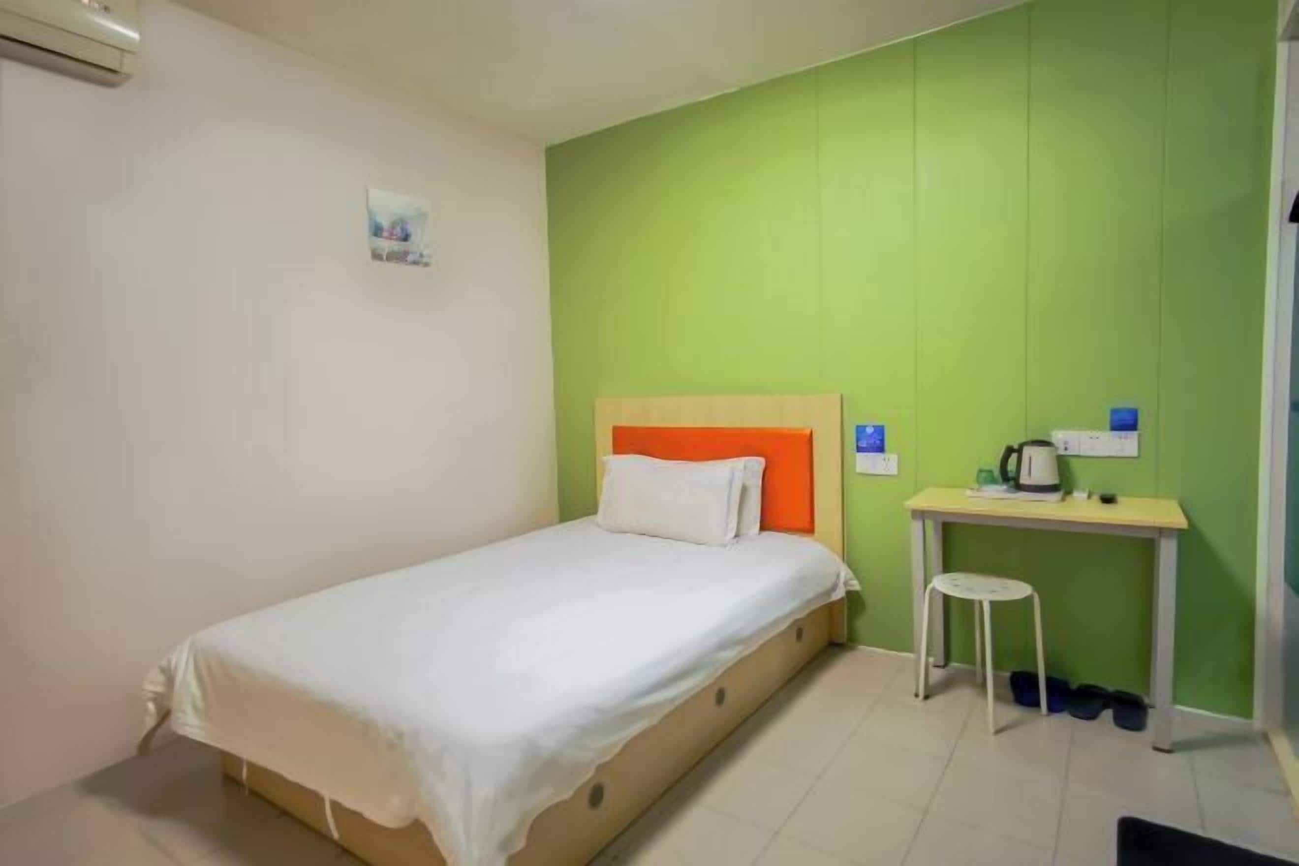 A single bed with a white mattress and an orange headboard, a small table with a chair, and a wall-mounted air conditioning unit.