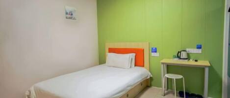 Una cama individual con colchón blanco y cabecera naranja, una mesita con una silla, y un aire acondicionado de pared.