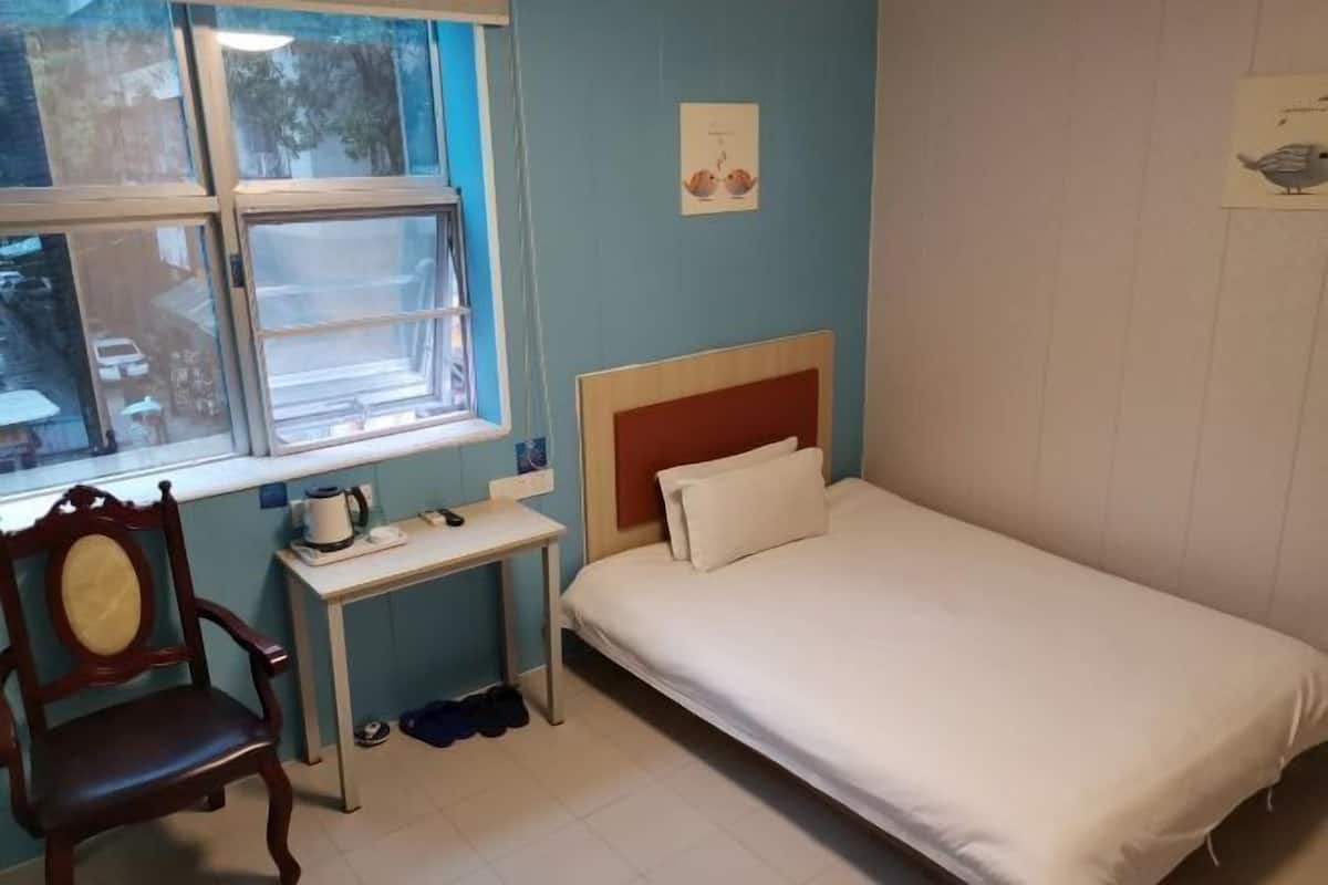 Quarto com uma cama de solteiro, uma cadeira de madeira, uma mesinha com chaleira e uma janela com vista para a rua.