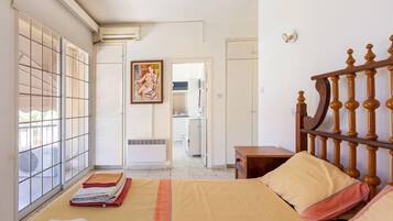 Un dormitorio con cama, cabecera de madera, ventana con persianas, un cuadro en la pared y un baño visible a través de una puerta abierta.